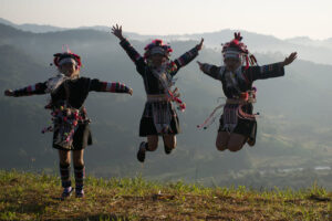 Tres mujeres de la etnia Akha, vestidas con trajes tradicionales coloridos y detallados, saltan de alegría en una colina al atardecer, mostrando la riqueza cultural de las comunidades locales en China