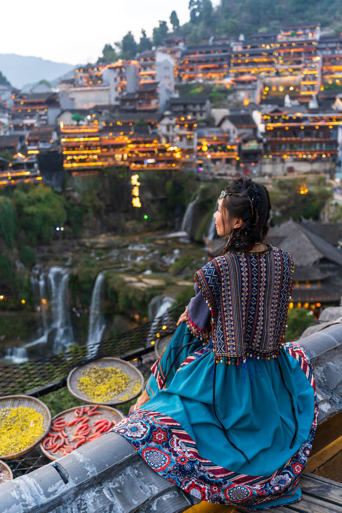 Mujer con vestimenta tradicional china sentada sobre un tejado antiguo, contemplando la vista panorámica de un pueblo histórico iluminado al atardecer, con cascadas de fondo y bandejas de secado de especias en primer plano