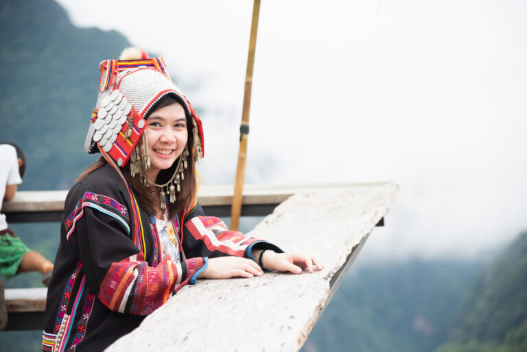 Joven sonriente vistiendo ropa y tocado tradicional de una minoría étnica, apoyada en una estructura de madera con un paisaje montañoso brumoso de fondo en China